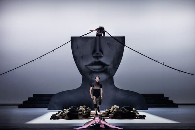 A stage set with a giant sculpted face, a dancer walking forward past dancers lying on the floor