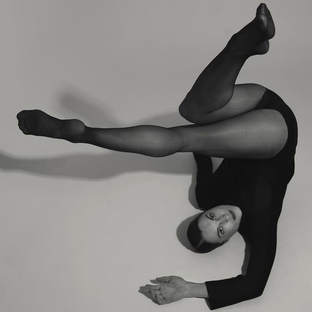 A dancer in black lies on a pale floor, legs folded overhead, looking up at the camera